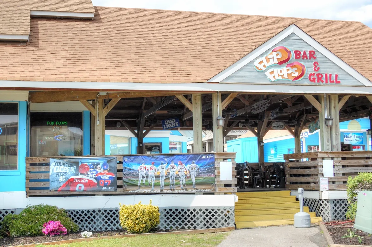 An exterior shot featuring the colorful signage of 'Flip Flops Bar & Grill', vibrant banners fluttering above, and a lively outdoor seating area ready for guests to dine and socialize. The atmosphere is relaxed and inviting, with cues to local fun and casual gatherings just steps from the property.