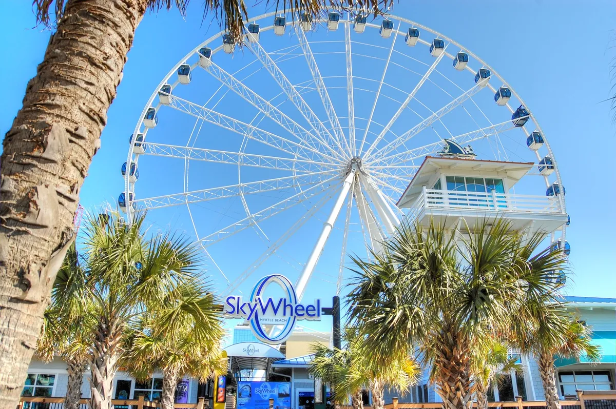A vibrant exterior image featuring the iconic Myrtle Beach SkyWheel, palm trees, and the entrance sign. The SkyWheel looms large in the background, offering a sense of excitement and adventure, while visitors can be imagined enjoying the view and taking memorable photos.