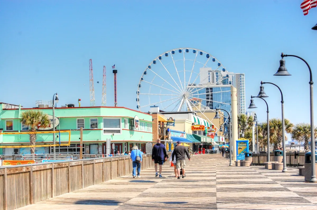 A lively boardwalk scene featuring a ferris wheel, colorful buildings, and people enjoying themselves as they explore shops and amusement rides. The mood is joyful and nostalgic, with the energy of Myrtle Beach's iconic attractions in full view.