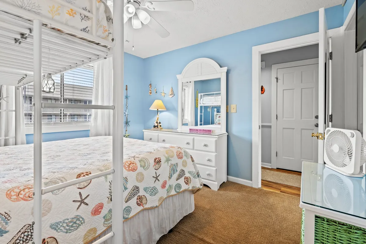 The image displays another angle of a secondary bedroom, featuring a bunk bed, a dresser, a mirror, a lamp, and decorative sea-themed items. Soft sunlight creates a cozy, inviting ambiance, suggesting a space that's both functional (ample storage) and charming for children or guests.