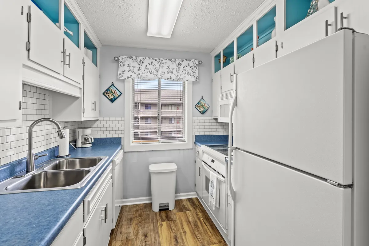 Here, the kitchen is in focus, with a gleaming sink, refrigerator, ample countertop space, and a window with curtains letting in cheerful daylight. The scene radiates a practical yet inviting atmosphere, perfect for preparing family feasts or snacks.