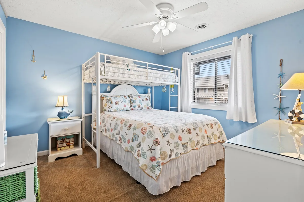 The second bedroom is shown with a bunk bed and another bed dressed in decorative bedding. The bright blue walls, playful accents, and window allowing natural light create a cheerful yet cozy vibe, perfect for families or friends. A nightstand with a lamp enhances the bedtime routine feel.