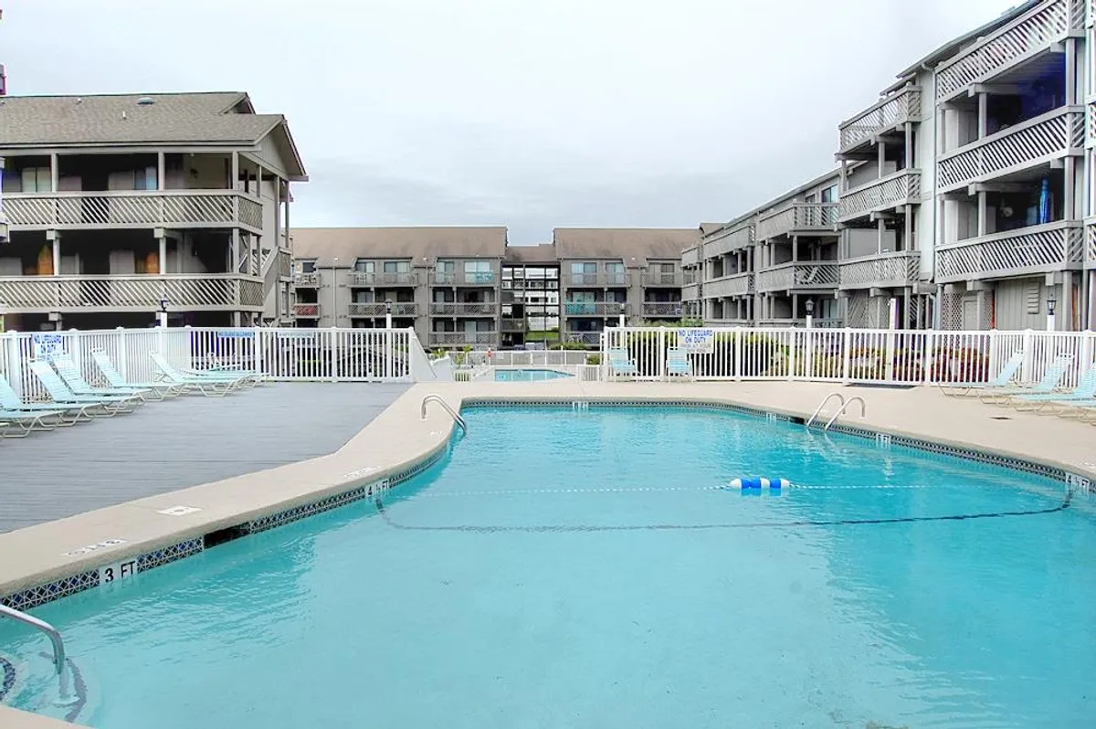 A bright, casual poolside scene with a clear swimming pool bordered by comfortable lounge chairs and the surrounding condo buildings. The relaxed setting is ideal for sunbathing, swimming, or enjoying poolside meals. The environment suggests a friendly, welcoming vibe.