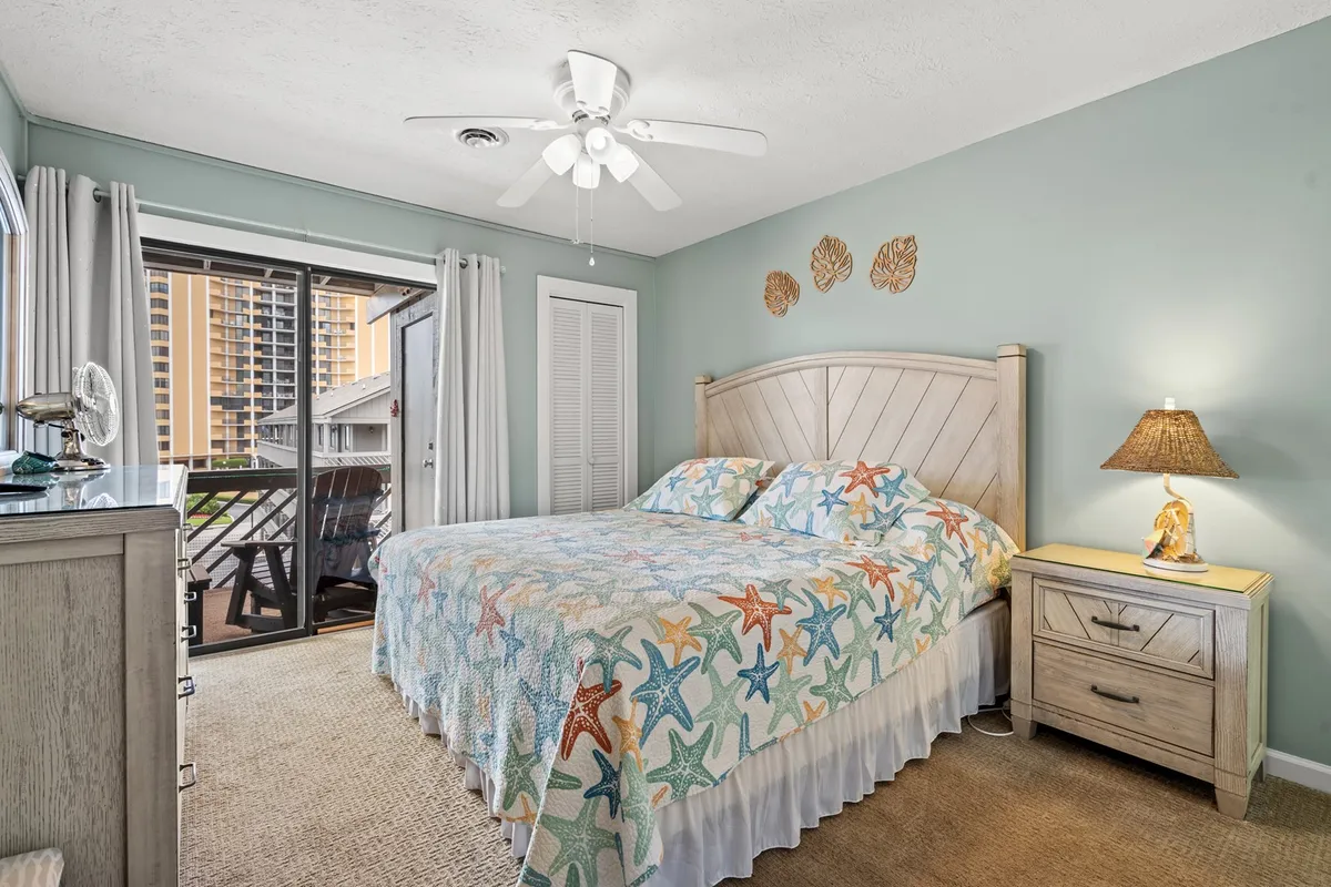 A tranquil bedroom featuring a bed with a starfish-patterned quilt, a nightstand with a lamp, and a sliding glass door that leads directly to the balcony. Soft natural light filters into the room, hinting at the inviting outdoor space just steps away.