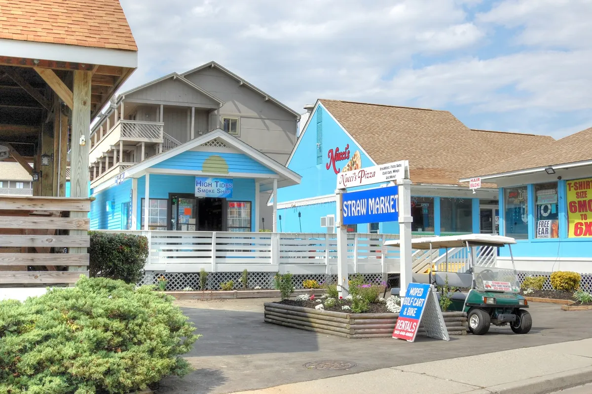 A bustling corner with the Straw Market sign, Nina's Pizza restaurant, and colorful blue storefronts, alive with people shopping, dining, and enjoying the community's sunny vibe. The scene is lively yet relaxed, hinting at local flavor and easy access to shopping and dining.
