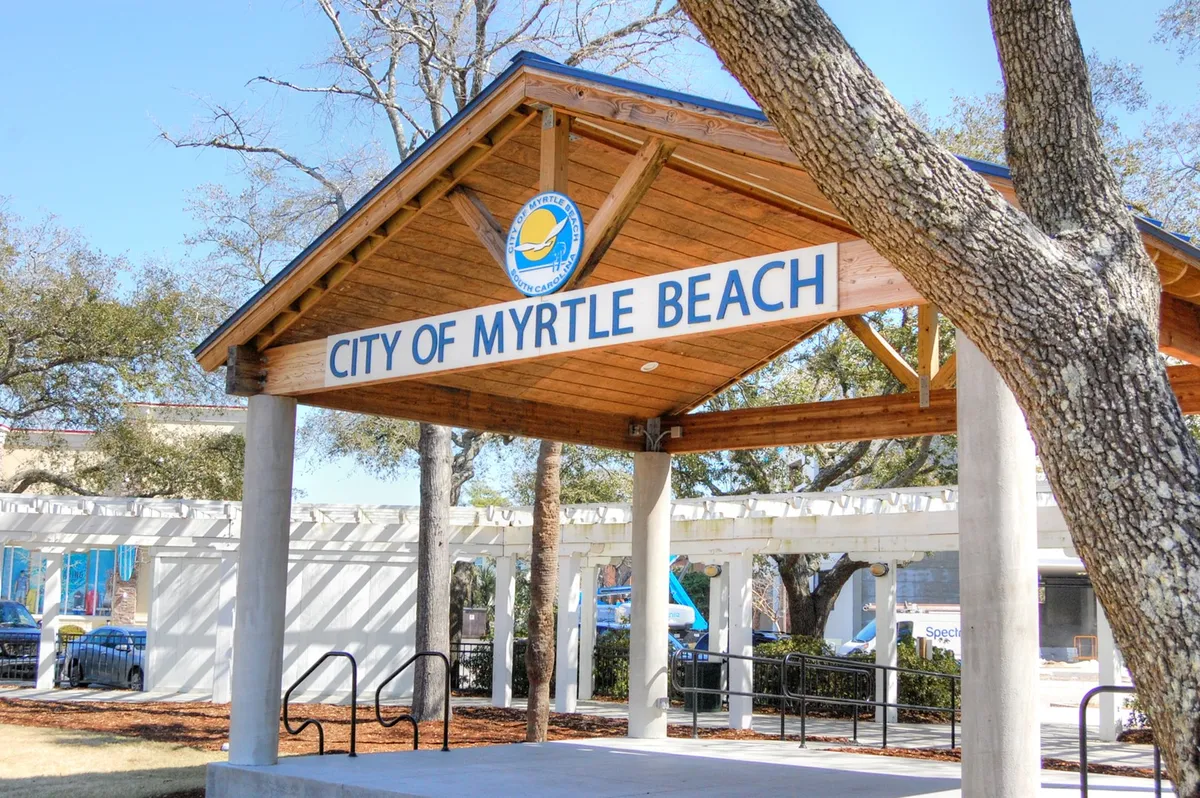 An inviting exterior image of the City of Myrtle Beach sign, set beneath a wooden pavilion and surrounded by trees. The setting suggests community involvement, upcoming events, and the lively spirit of the destination.