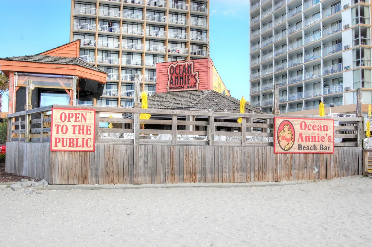 The image captures the vibrant exterior of Ocean Annie's Beach Bar, located right by the beach. Yellow umbrellas, outdoor bar seating, and casual fencing create a lively, social atmosphere. The bar is a hub for guests to enjoy drinks, live music, and the ocean breeze, as described in the section’s emphasis on resort amenities and social spaces.