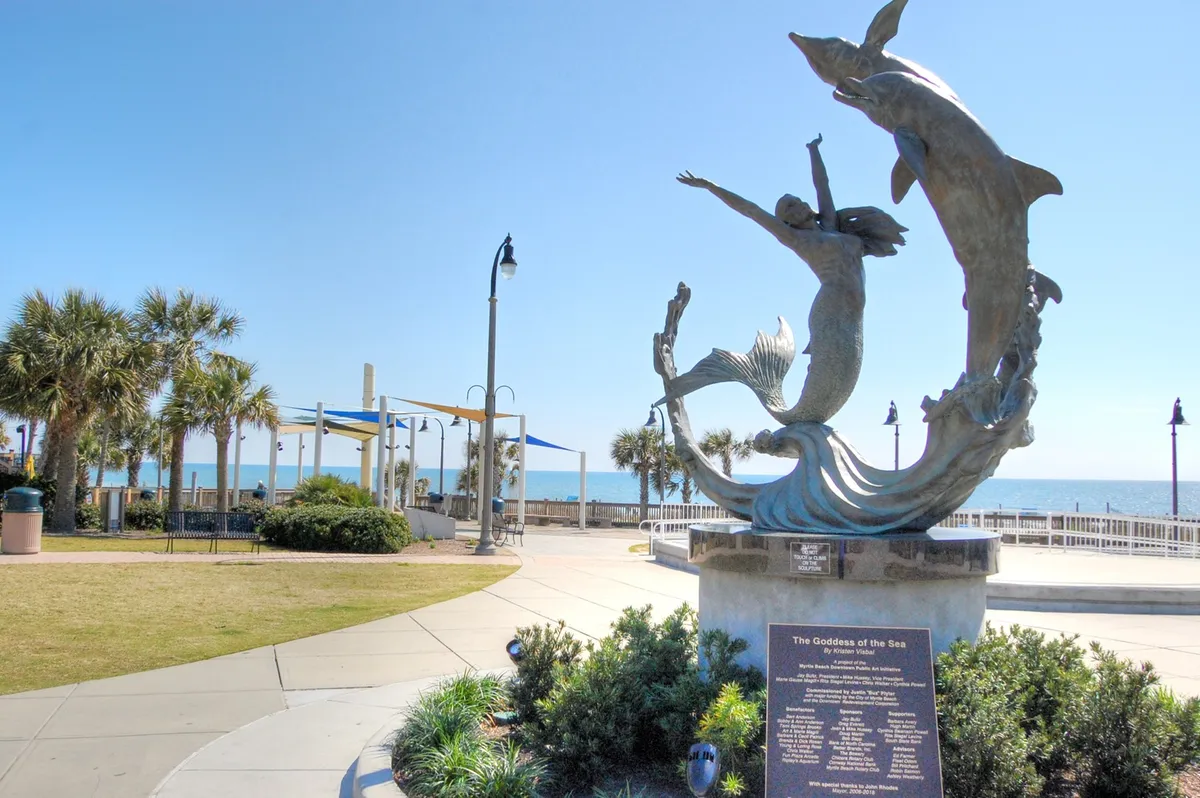 A wide exterior view showing the iconic Goddess of the Sea sculpture set against the backdrop of the ocean, framed by palm trees. The scene captures the spirit of Myrtle Beach—welcoming, scenic, and rich with opportunities for walks, seaside relaxation, and memorable photo moments.
