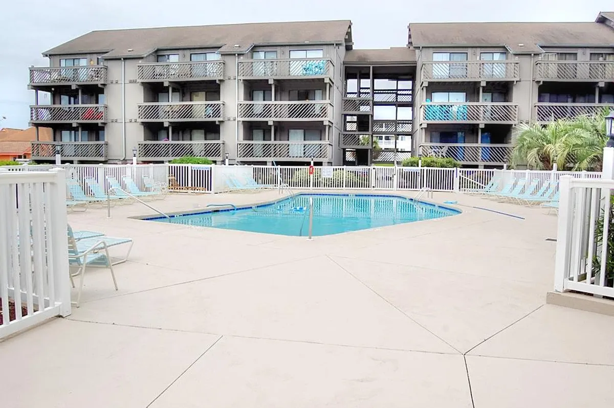 An inviting outdoor swimming pool surrounded by lounge chairs and the balconies of the condo building. People are relaxing by the water and socializing, creating a lively yet laid-back atmosphere. The pool area is spacious with ample seating, perfect for group gatherings or quiet sunbathing.