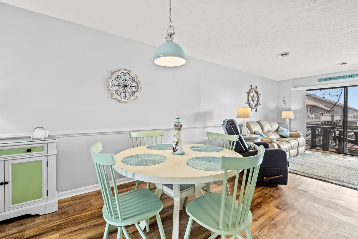 The photo shows a stylish dining area with a wooden table surrounded by light blue chairs, a decorative wall clock, a modern light fixture, and a sliding glass door leading to an outdoor deck, allowing natural light to fill the space. The arrangement highlights the transition between indoor and outdoor living and the potential for memorable shared meals.
