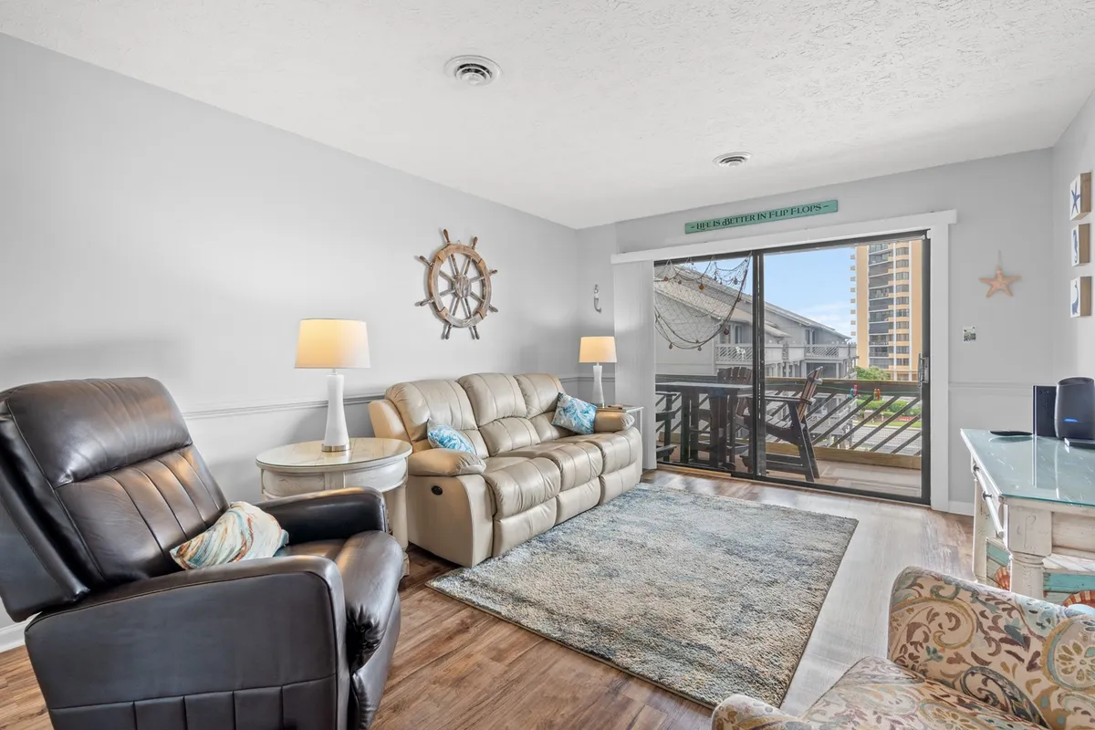The hero image showcases the condo's inviting living area, featuring a plush sofa and recliner set against a large window that offers a serene view. Nautical touches like a decorative ship wheel and starfish create a beachy, coastal vibe. The scene is bathed in natural light, hinting at beautiful sunsets and a relaxing atmosphere perfect for unwinding after a day at the beach.