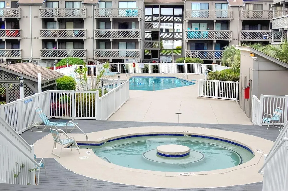 A wide exterior view showing the resort’s swimming pool, hot tub, lush greenery, and the balconies of the building above. The image conveys tranquility and offers a visual context for the balcony’s vantage point over the resort amenities.